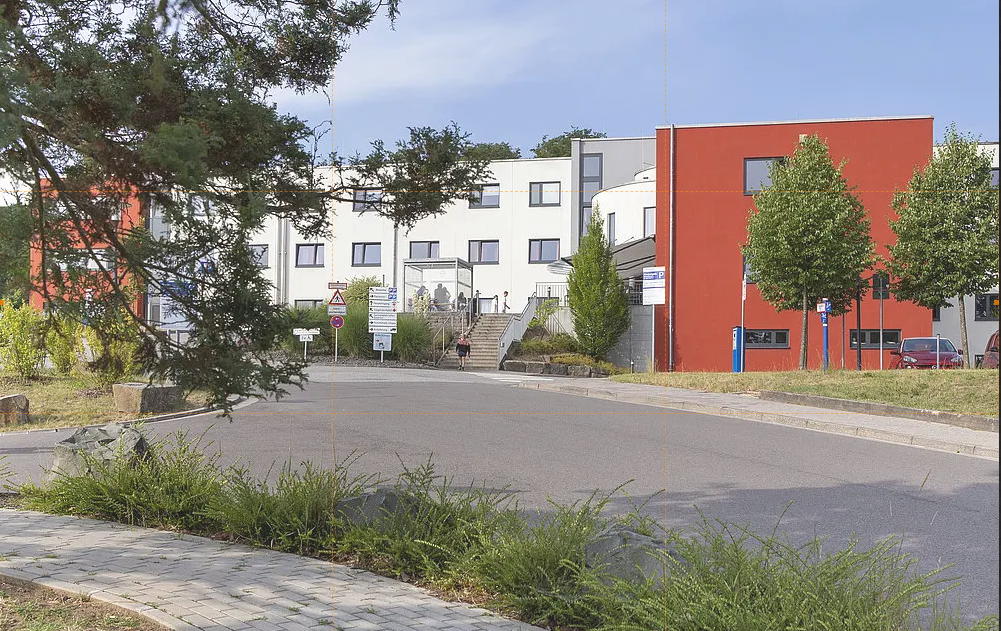 Gebäudekomplex mit weißer und roter Fassade hinter einer Straße und Bäumen im Vordergrund
